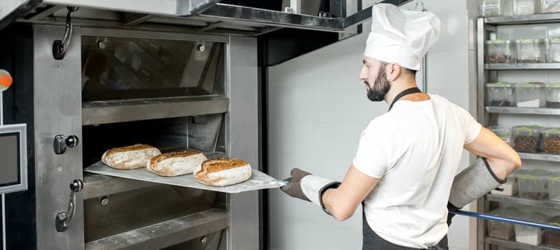 Baker taking off baked breads with shovel from the professional oven at the manufacturing