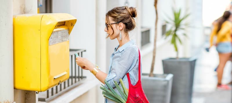 Young woman putting letter to the old yellow mailbox standing with mesh bag full of food outdoors on the street