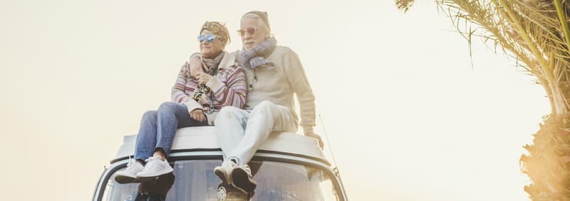 Wanderlust and travel destination happiness concept with old senior beautiful couple sitting and enjoying the outdoor freedom on the roof of vintage van vehicle together - sun backlight