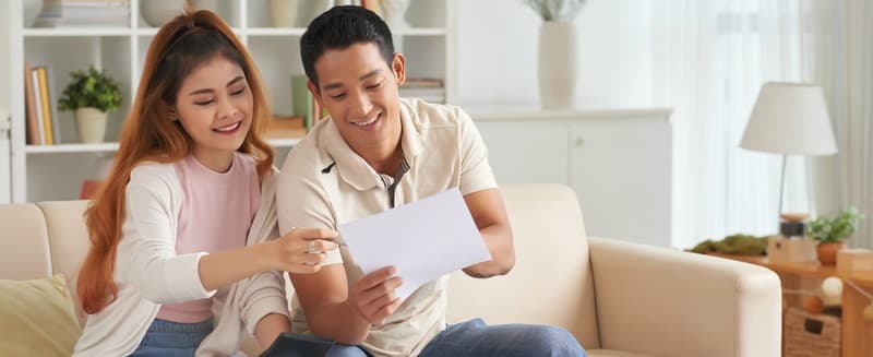Portrait of young Asian couple planning family budget in new house, smiling and talking sitting on sofa