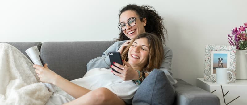 Two happy young casual women having fun using smartphone at home. Lesbian millennial couple.